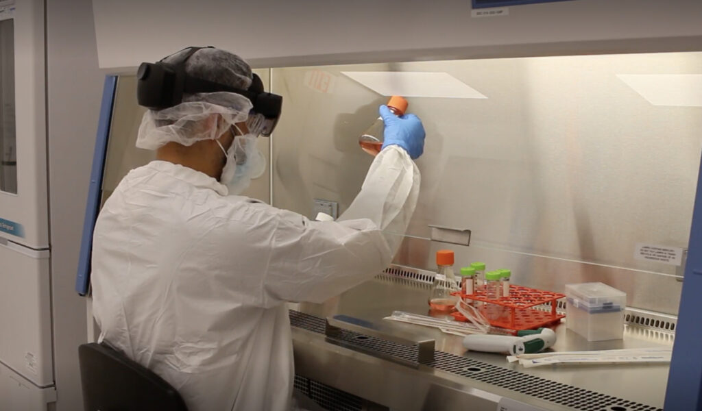 Man wearing gown and Microsoft HoloLens works in cleanroom and holds up a flash with orange liquid.
